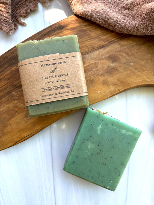 A bar of green goat milk soap with exfoliants, labeled 'Desert Dreams', resting on a wooden surface.